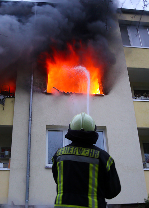 Flammen schlagen aus der Wohnung eines Mehrfamilienhauses. Ein Feuerwehrmann löscht. Foto: Stefan Rampfel (rammi76 Adobe Stock)